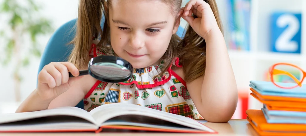 Young girl with pigtails looking through a magnifying glass at an open book on a table.