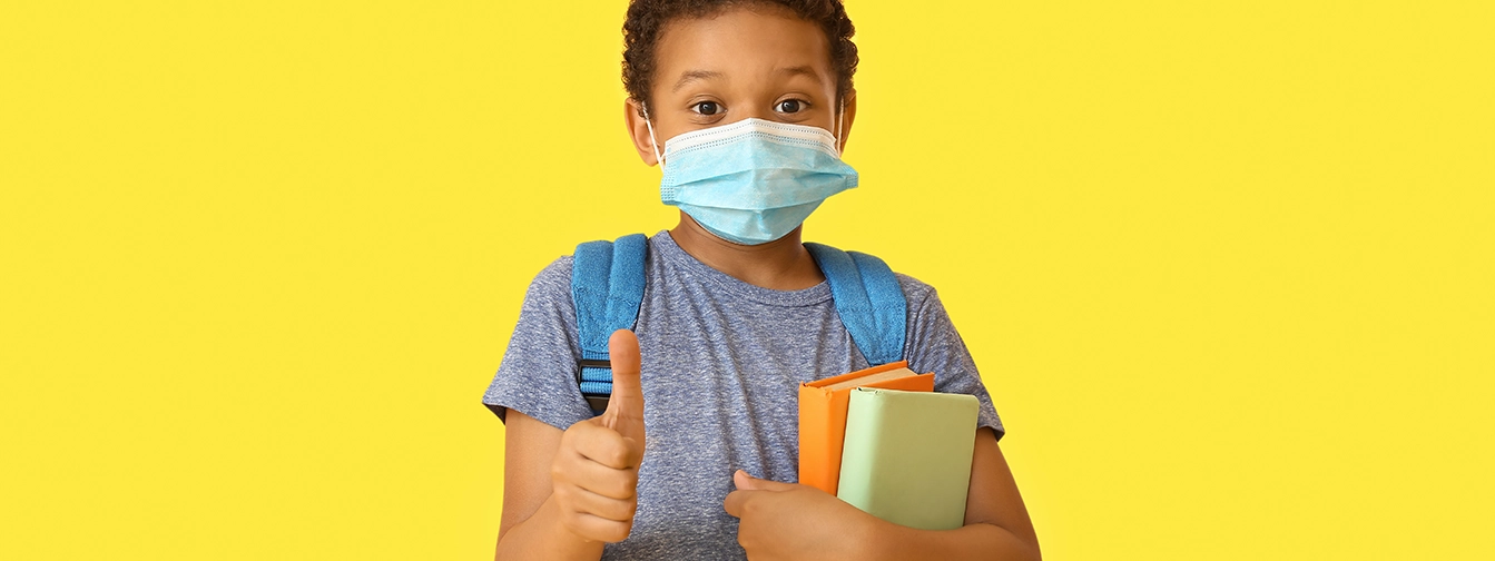 A young African-American boy in a backpack and a pandemic mask holds two colorful books under his left arm and makes the thumbs up gesture with his right hand.