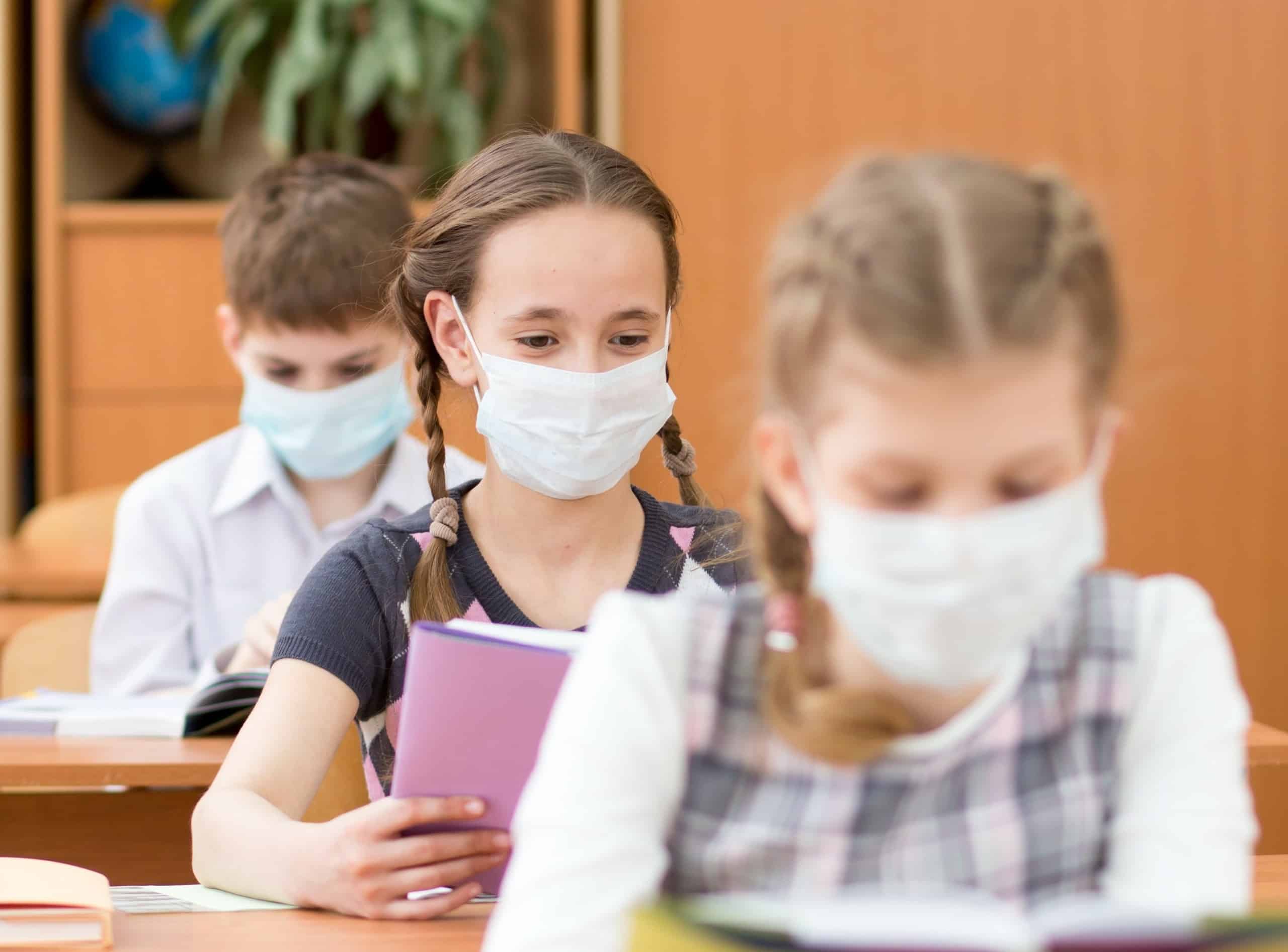 Schoolkids with medicine mask on faces against virus in classroom; keeping reading social while distancing